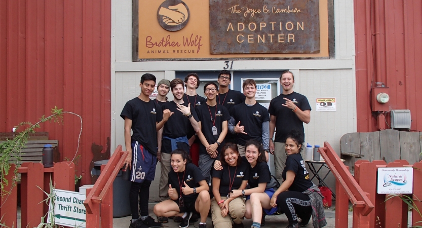 A group of students smile at the camera on the steps of a building under a sign. The sign reads "Brother Wolf Animal Rescue, Adoption Center." Additional text is not readable. 